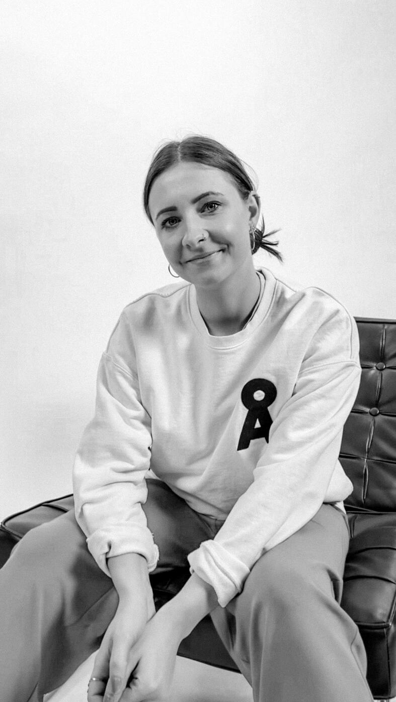 Schwarzweiß-Portrait von Almut, sitzt auf einem Sofa und trägt ein weißes Sweatshirt mit einer schwarzen 8