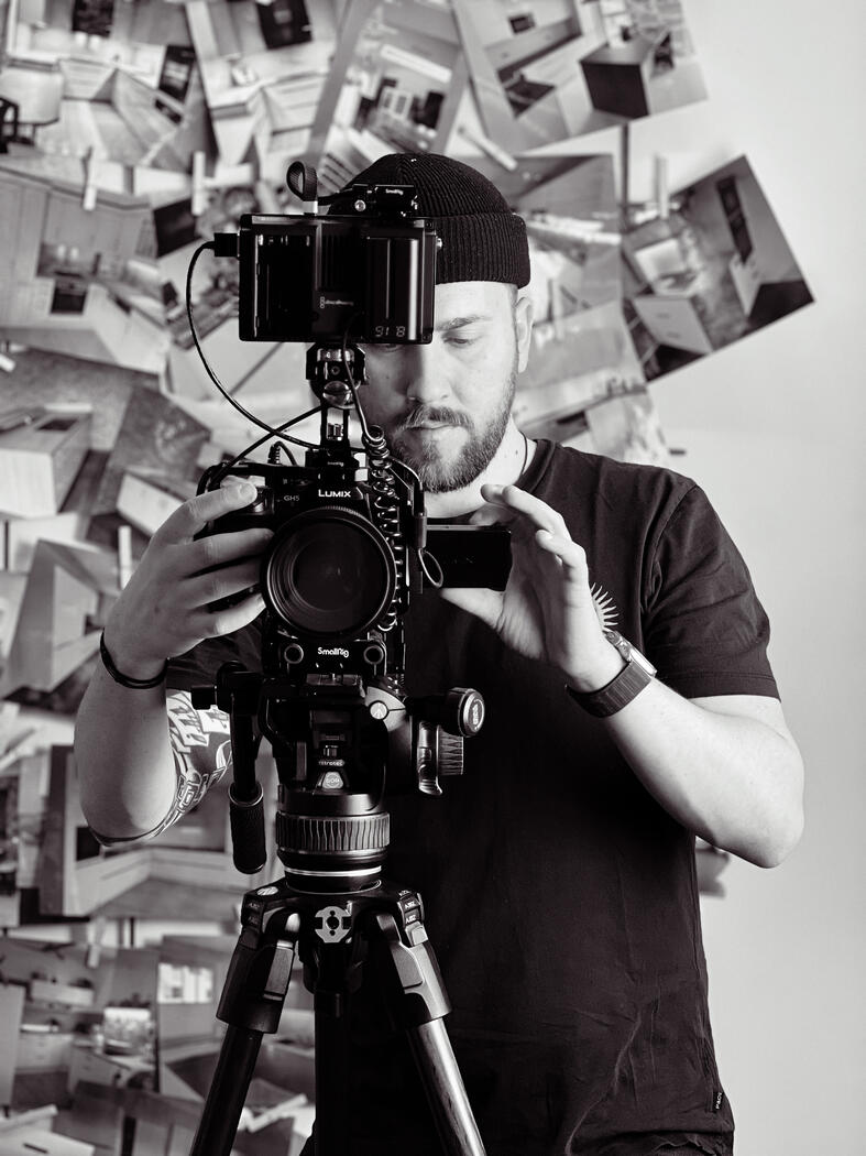 Schwarzweiß-Portrait von Robert mit Kamera in der Hand vor einer Fotowand